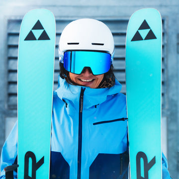 Ein lächelnder Skifahrer in blauer Ausrüstung und mit Helm trägt eine NAKED Optics Skibrille mit Anti-Fog-Beschichtung und hält leuchtend blaue Skier vor einer verschneiten, belüfteten Kulisse.