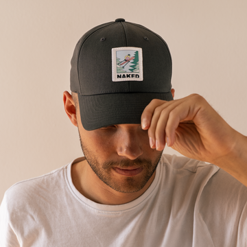 Ein Mann in einem weißen Hemd rückt eine dunkle NAKED Optics Unisex-Baumwoll-Baseballmütze mit einem Aufnäher zurecht, auf dem NAKED steht und eine Person beim Surfen abgebildet ist.