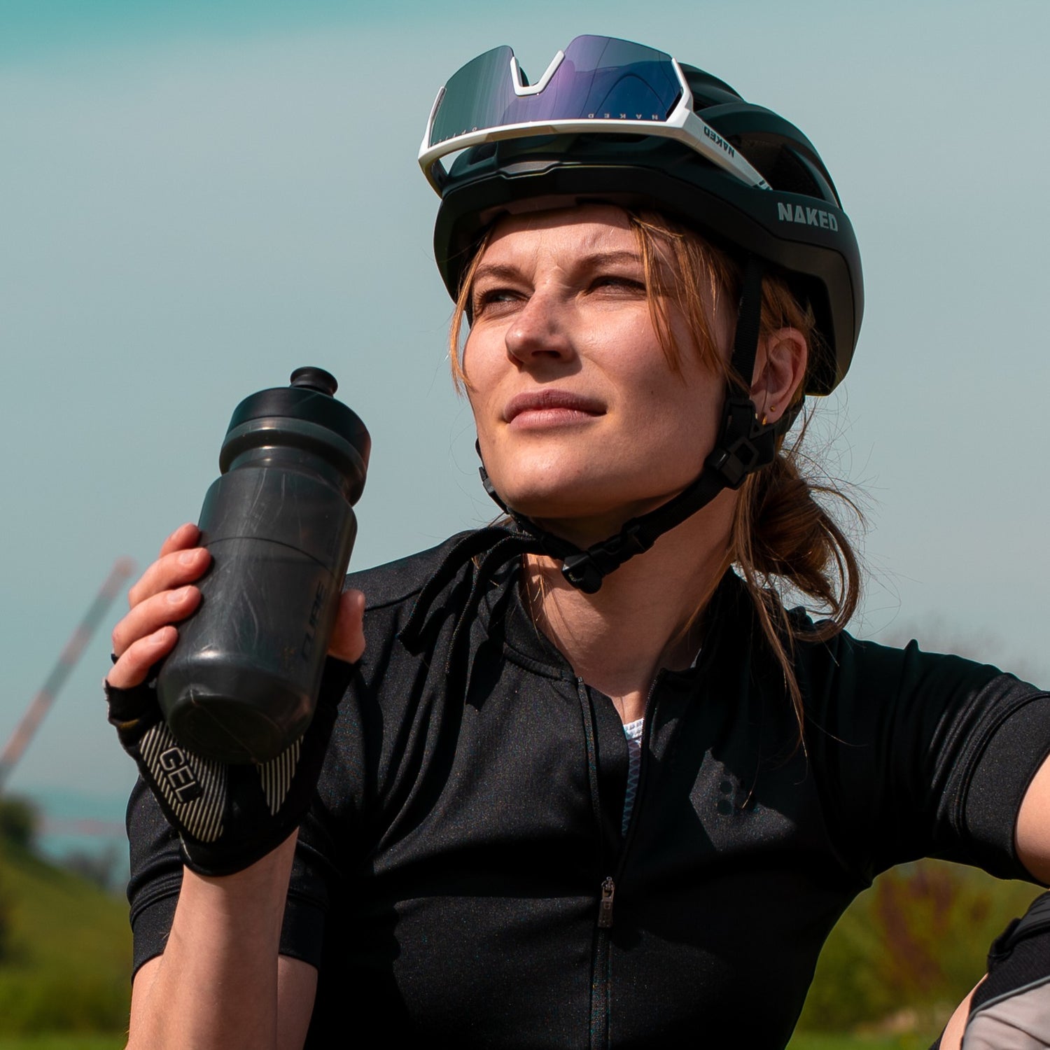 Eine Frau schaut in die Sonne und hebt ihre Trinkflasche an. Die trägt einen Rennradhelm und eine Fahrradbrille; Credits: Kathy Lerchner