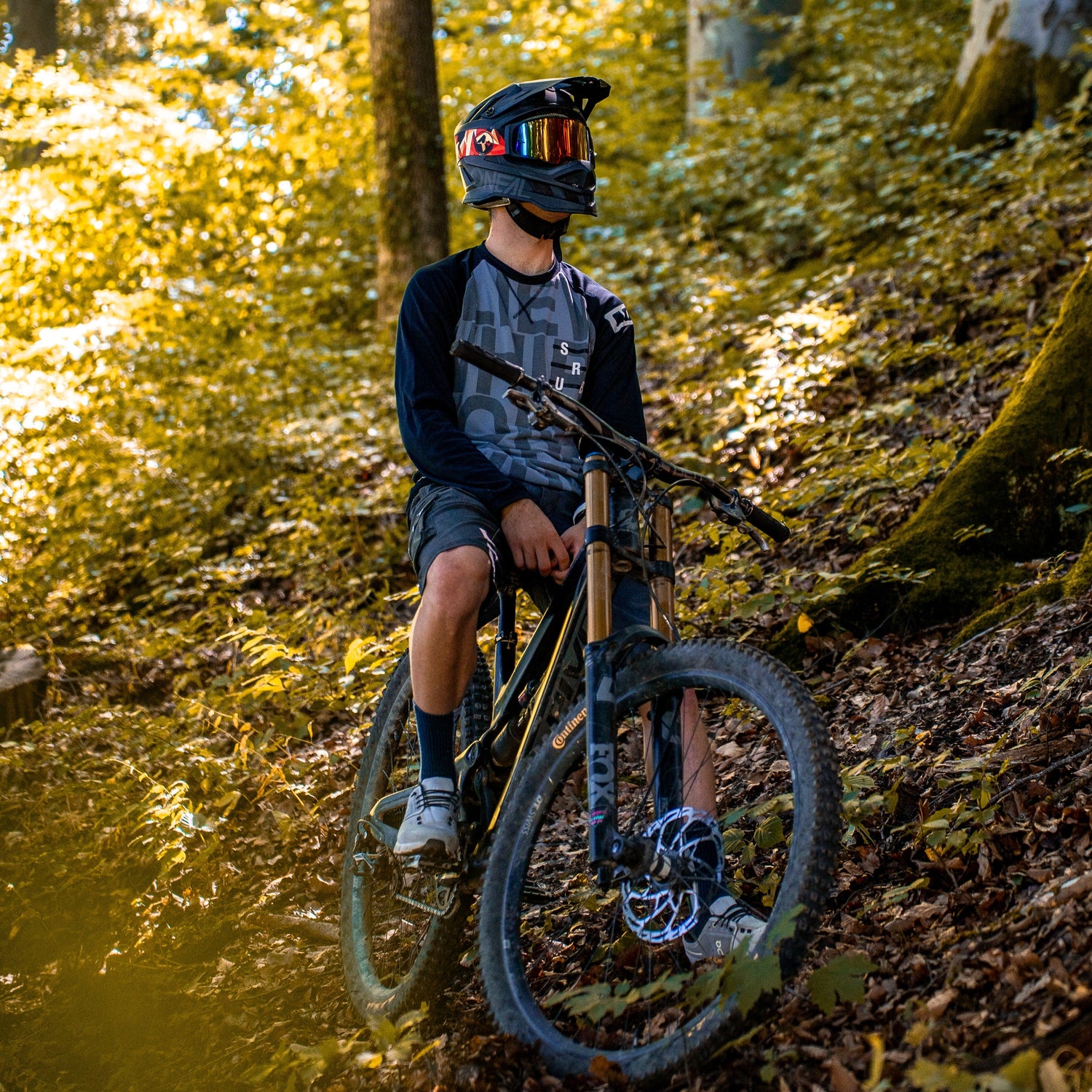 Ein Mann steht mit seinem Mountainbike im Wald auf einem Trail, er trägt einen Full-Face Helm mit einer verspiegelten Downhill Brille; Credits: Kathy Lerchner 