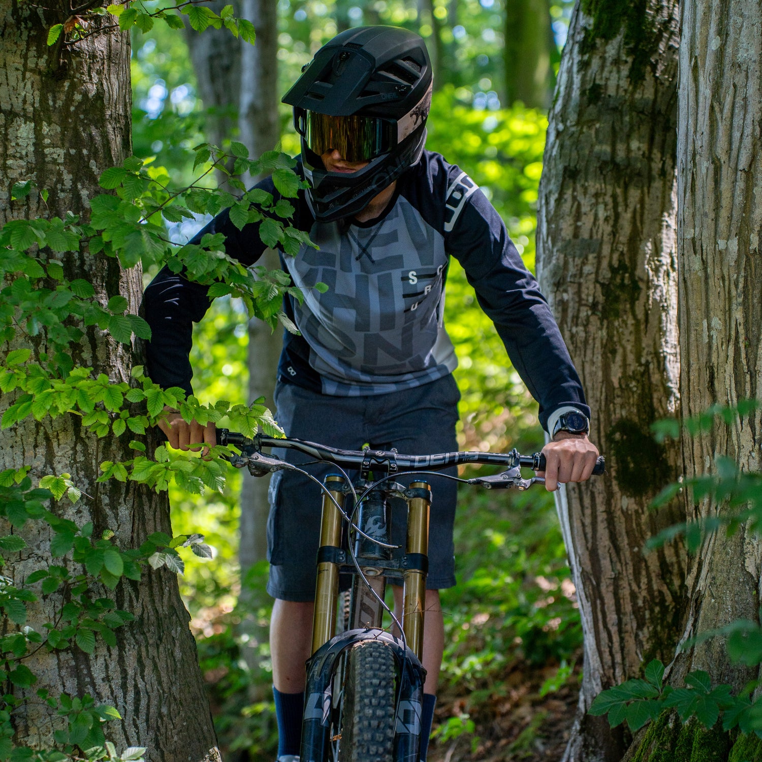 Ein Mann fährt mit seinem Mountainbike durch den Wald, er trägt einen Full-Face Helm mit einer auffälligen Downhill Brille; Credits: Kathy Lerchner