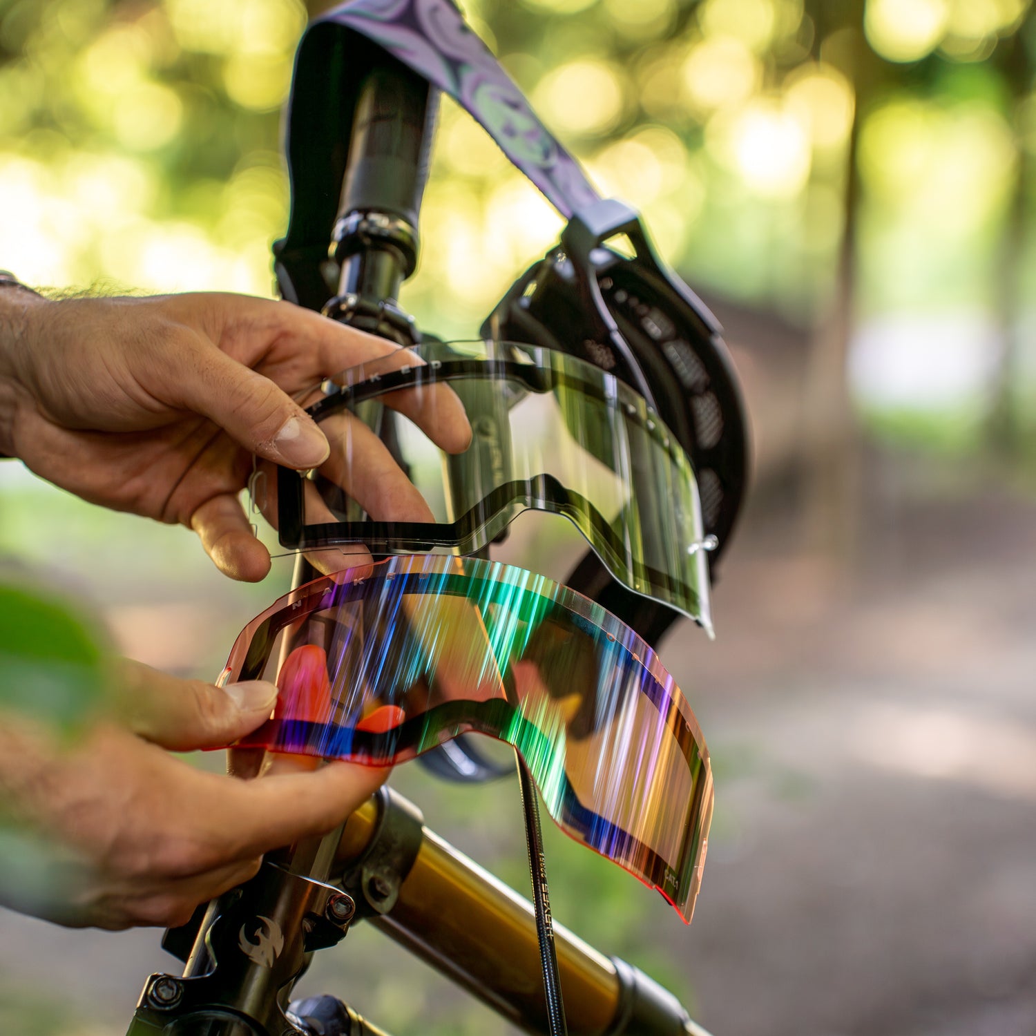 Hände, die zwei verschiedene NAKED Optics Downhillbrille-Gläser über einem Fahrrad halten - ein klares, ein farbiges. Verfügt über einen UV-400 Schutz für den Augenschutz und ein einfaches Magnetsystem Gläser für den einfachen Austausch von Gläsern.