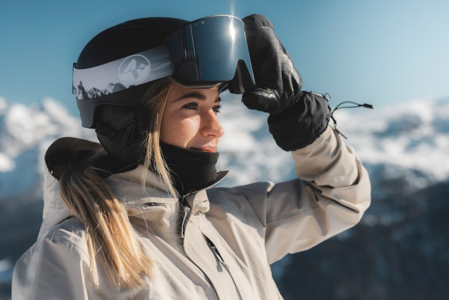 Eine blonde Skifahrerin steht in den Bergen und hebt ihre Skibrille vom Helm, um in die Sonne zu lachen; Credits: Mathäus Gartner