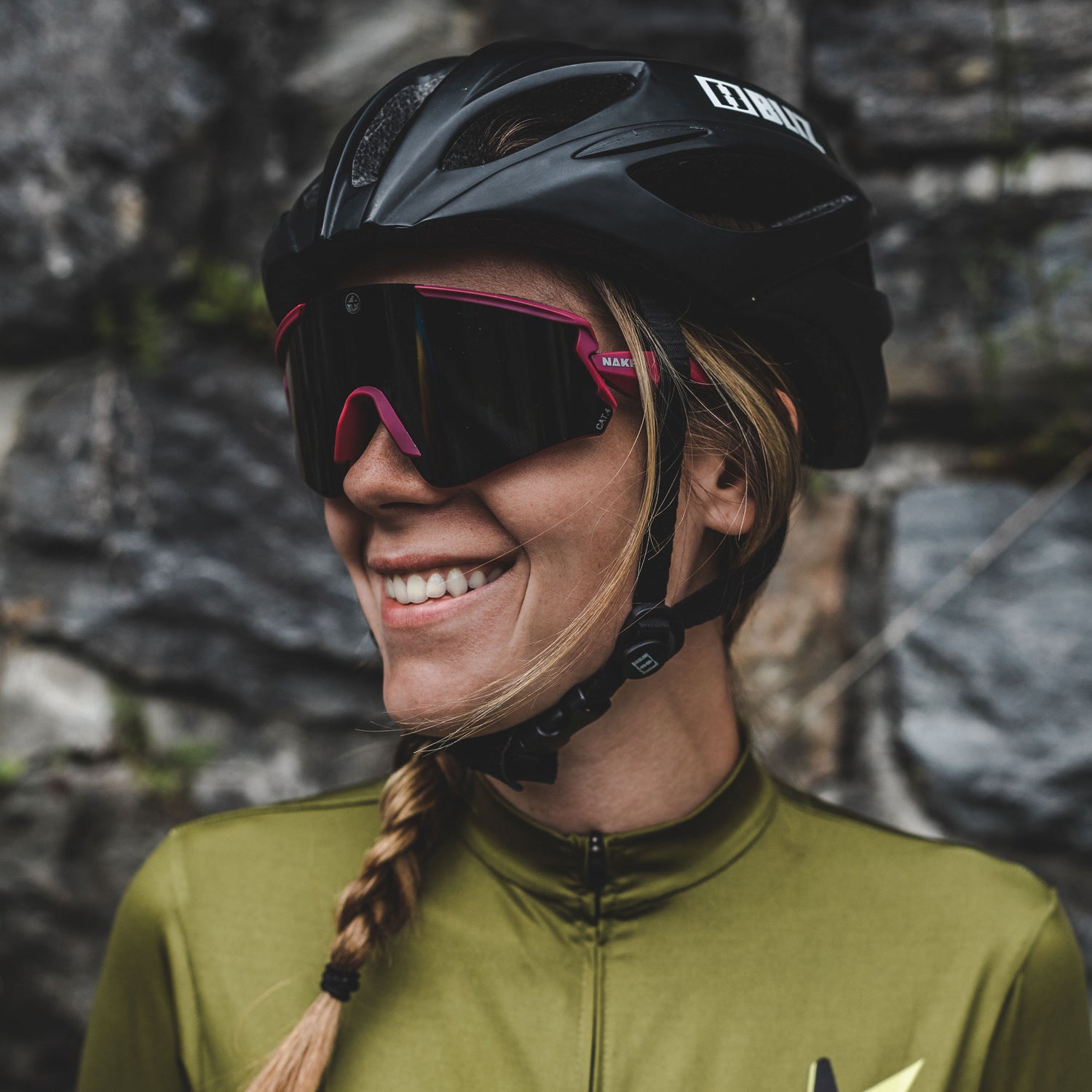Lächelnder Radfahrer mit Sonnenbrille und Helm von NAKED Optics, der ein grünes Trikot trägt und an einer Steinmauer steht - bereit für Outdoor-Abenteuer.