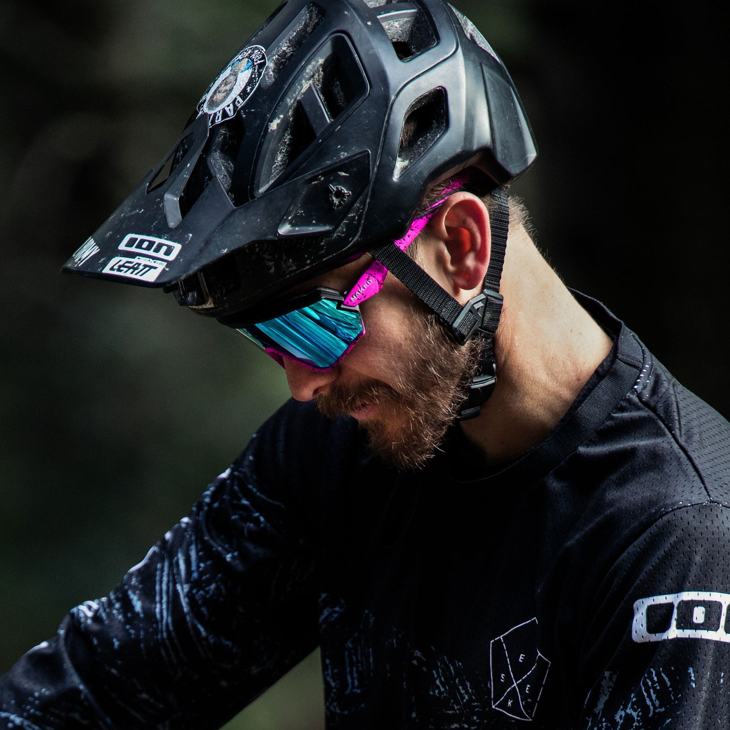 Eine bärtige Person trägt einen schwarzen Helm und eine reflektierende Sonnenbrille von NAKED Optics Fahrradbrille für kleine Köpfe und schaut im Freien nach unten.