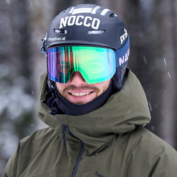 Eine Person mit Helm, grüner Skijacke und NAKED Optics Skibrille NOVA mit Anti-Beschlag-Beschichtung lächelt im Freien bei verschneitem Wetter.